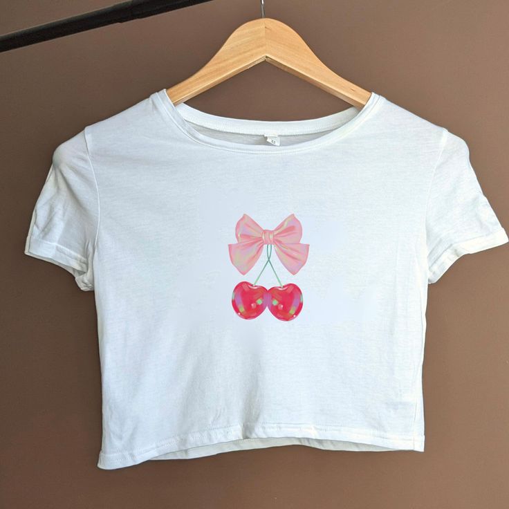 Introducing the Coquette Cherry Ribbon Bow Women's Crop Tee, a must-have addition to your wardrobe for those who appreciate softcore balletcore era fashion and trendy vintage cute aesthetic. This adorable t-shirt combines the elegance coquette with the charm of a holographic set of cherries with a ribbon bow. Made from high-quality materials, this crop tee offers both comfort and style. Embrace your inner princesscore art enthusiast and make a fashion statement with this versatile piece. Whether you pair it with high-waisted jeans for a casual yet chic look or with a skirt for a more polished ensemble, this crop tee is sure to turn heads. The soft fabric feels gentle against your skin, while the bow adds a touch of whimsy to your outfit. The Coquette Cherry Ribbon Bow Women's Crop Tee is p Aesthetic Graphic Print Tops For Summer, Cute White Cotton Cropped T-shirt, White Cotton Aesthetic T-shirt, Cute Summer Crop Top With Funny Print, Aesthetic Graphic Print Tops For Spring, Cute Summer Crop Top T-shirt, Summer Aesthetic Short Sleeve T-shirt, White Aesthetic Summer Tops, Summer Cotton Cropped T-shirt With Funny Print