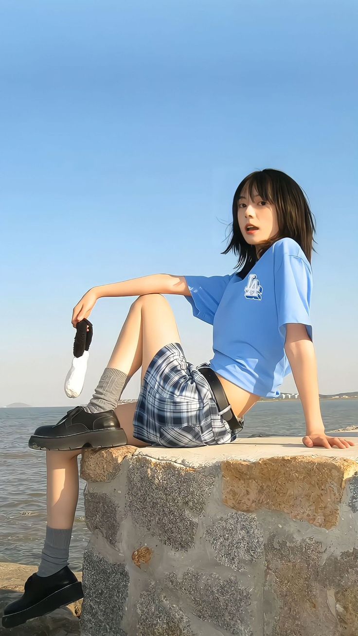 a young woman sitting on top of a stone wall next to the ocean with her legs crossed