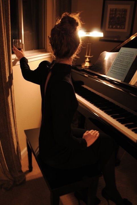 a woman sitting at a piano with a glass of wine in her hand and looking out the window