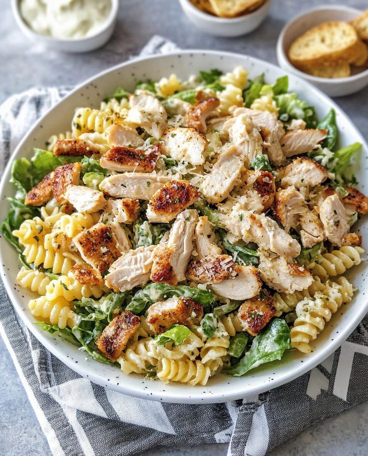 a white bowl filled with pasta and chicken on top of a blue table cloth next to bowls of bread