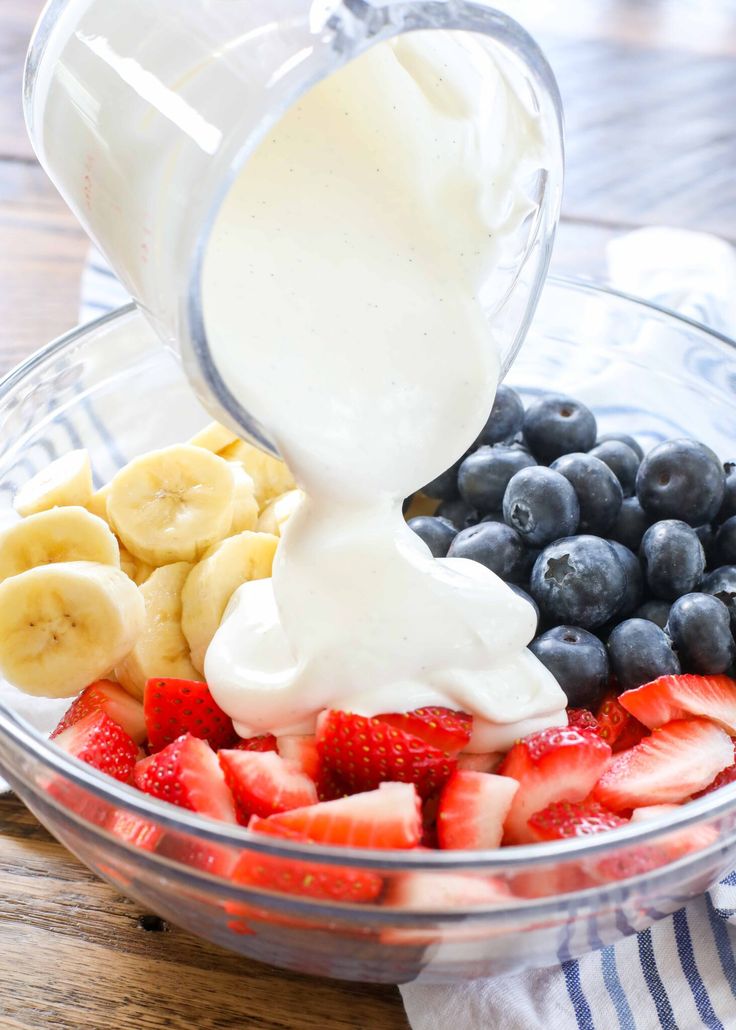 fruit and yogurt being poured into a bowl with blueberries, strawberries, bananas, and more