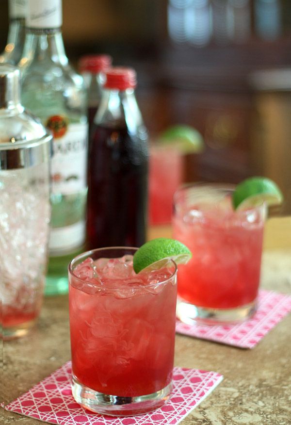 two cocktails with lime garnish sit on napkins next to liquor bottles