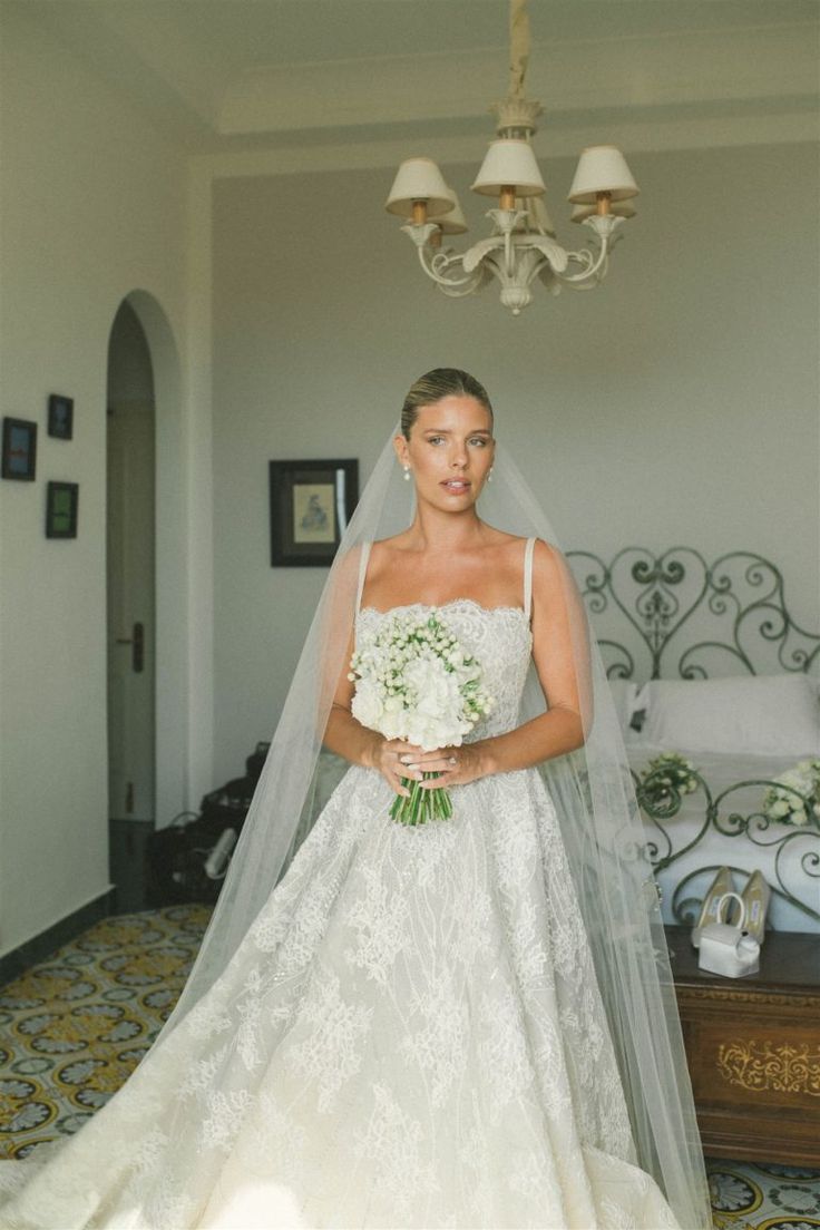 a woman in a white wedding dress standing next to a chandelier and holding a bouquet