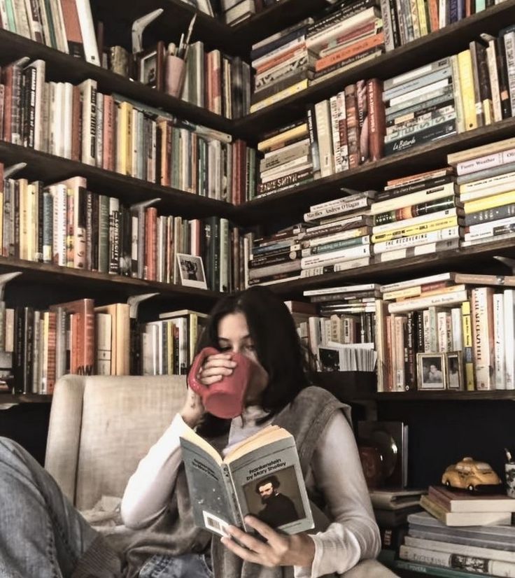 a woman sitting in a chair holding a book and drinking from a cup while looking at the camera