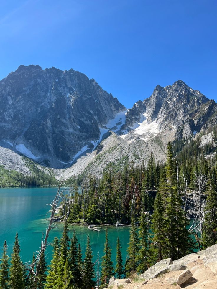 a mountain lake surrounded by pine trees and snow covered mountains in the distance with blue sky