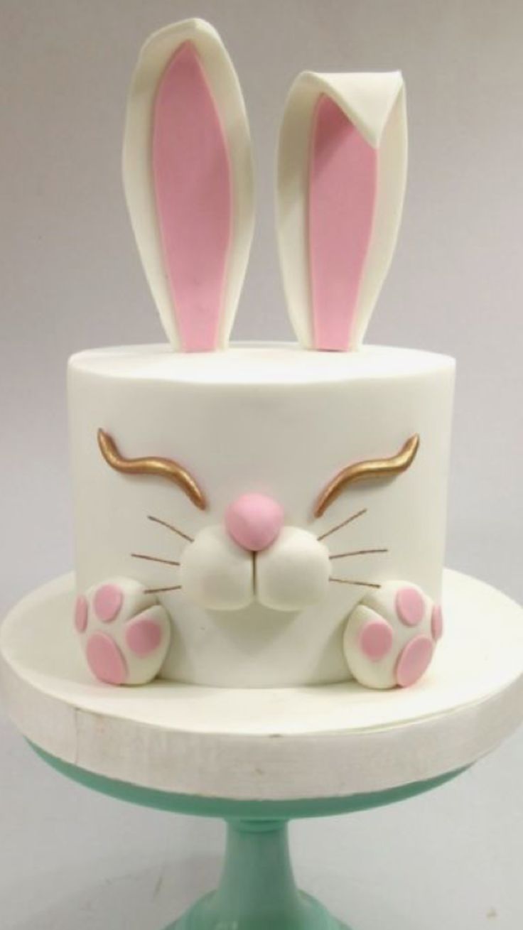 a white cake with pink and gold bunny ears on it's face, sitting on top of a green pedestal