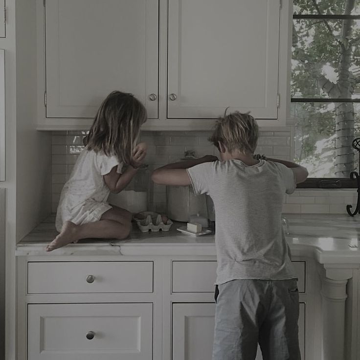 two children in the kitchen playing with each other's hair and looking out the window