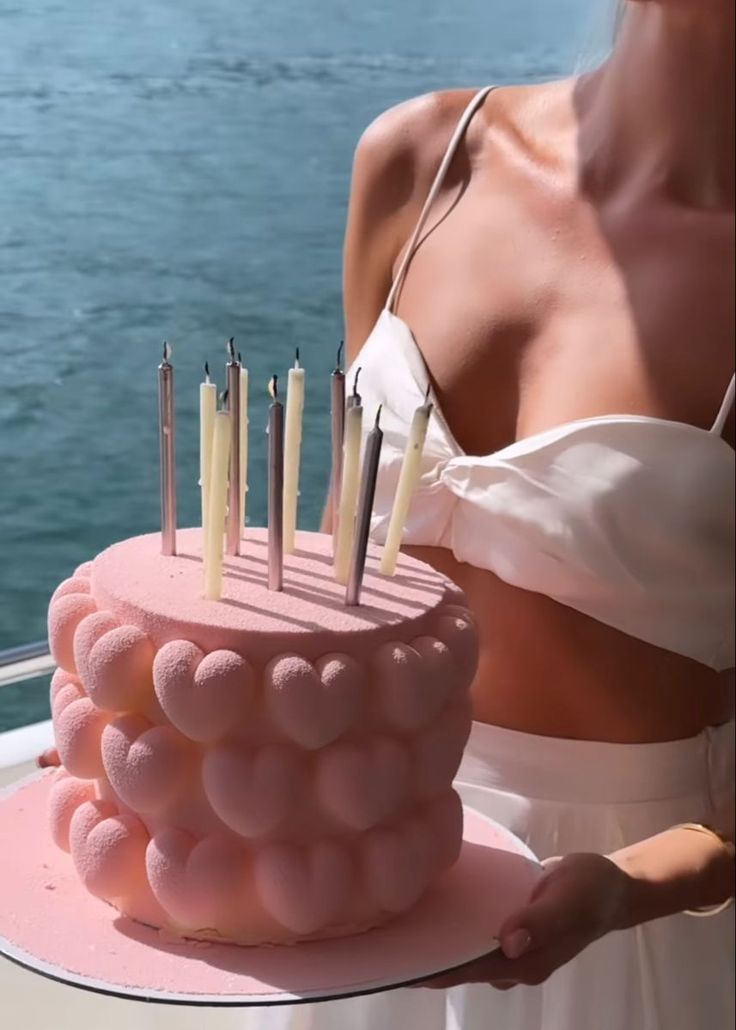 a woman is holding a pink cake with candles on it while standing next to the water