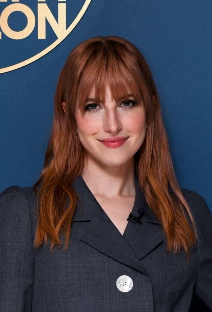 a woman with red hair wearing a suit and smiling at the camera while standing in front of a blue wall