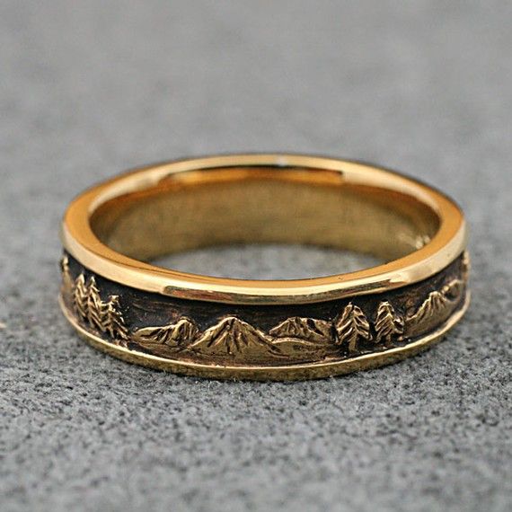 a gold wedding band with trees and mountains on it, sitting on the ground in front of a gray background