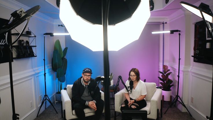 a man and woman sitting in chairs next to each other with lighting equipment behind them