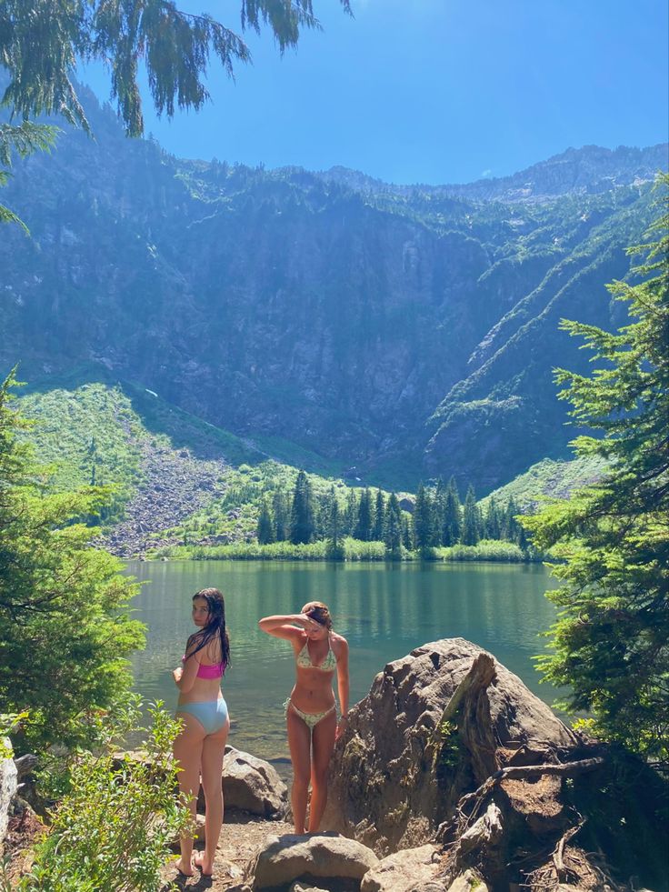 two women in bikinis are standing on rocks near a lake with mountains in the background