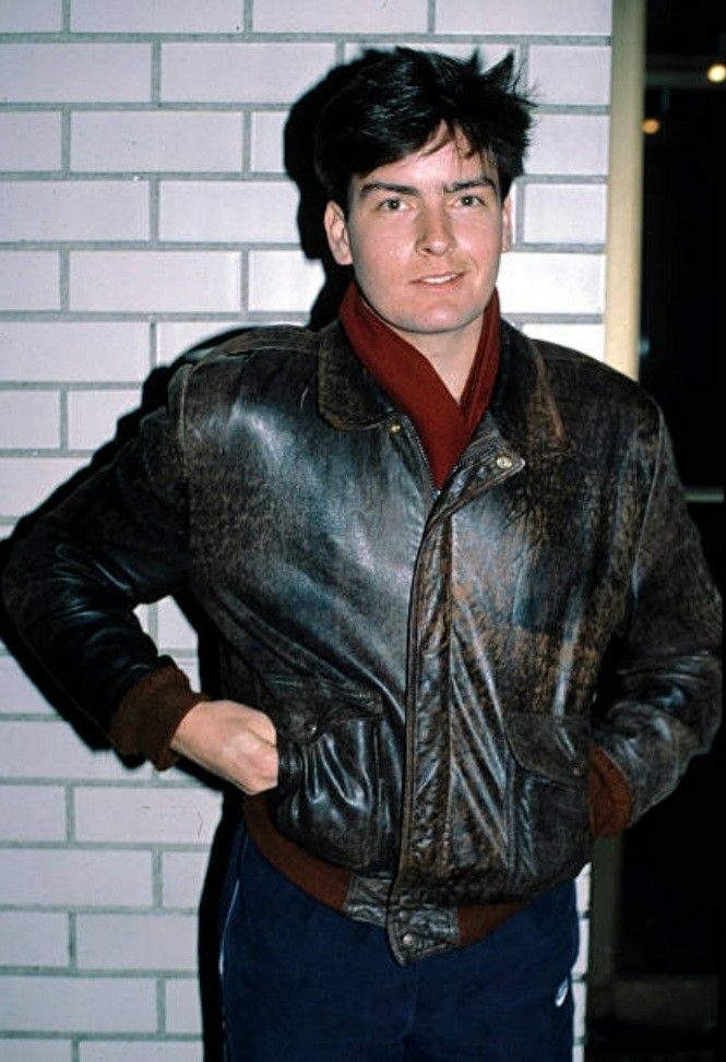 a man wearing a leather jacket standing in front of a white brick wall with his hands on his hips