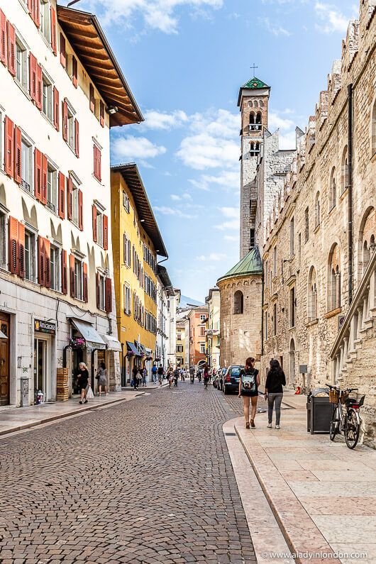 people are walking down the cobblestone street in an old european town with tall buildings