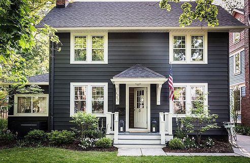 a black house with white trim and two windows on the second floor, has an american flag at the front door