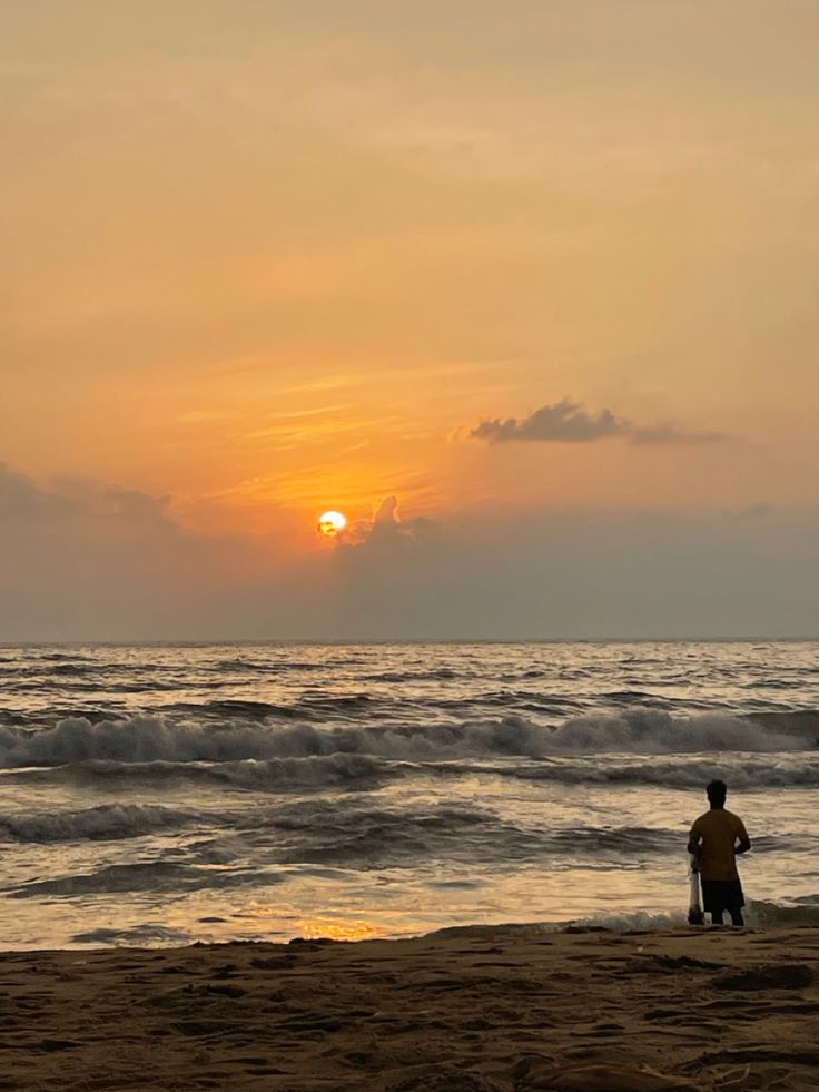 a man standing on top of a sandy beach next to the ocean at sunset or dawn