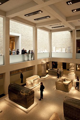 people looking at artifacts in a museum with lights on the ceiling and large windows above them