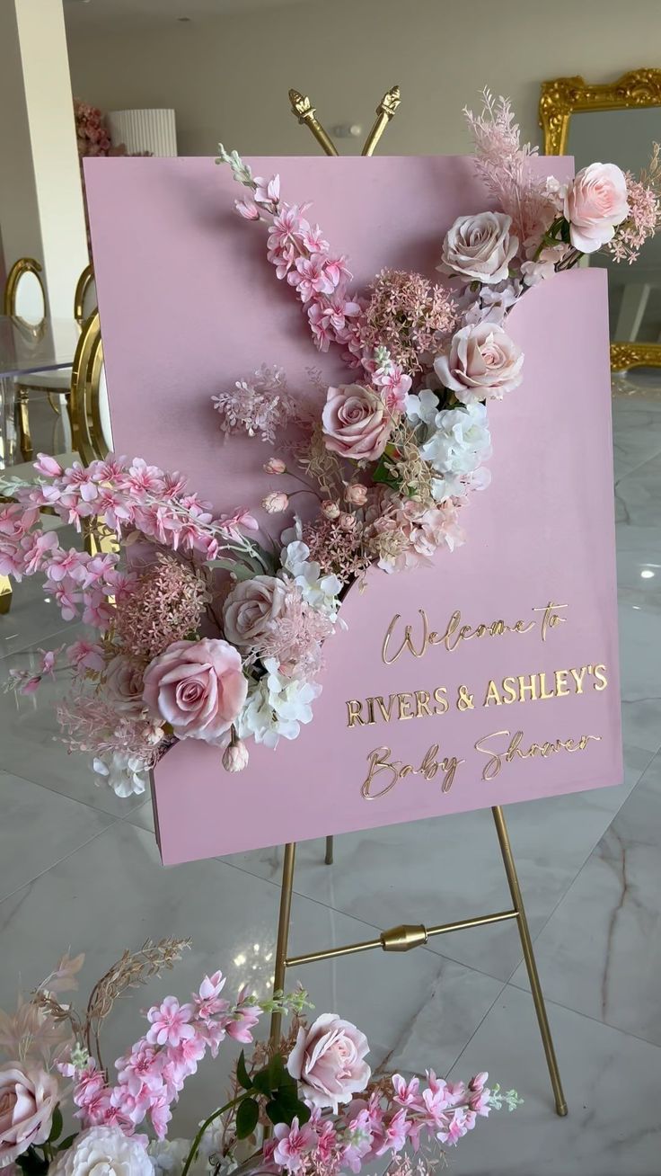 a welcome sign with pink flowers and greenery on it in front of a gold frame