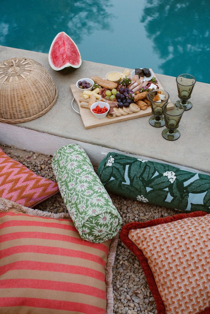 an assortment of food sitting on top of a table next to a pool with watermelon