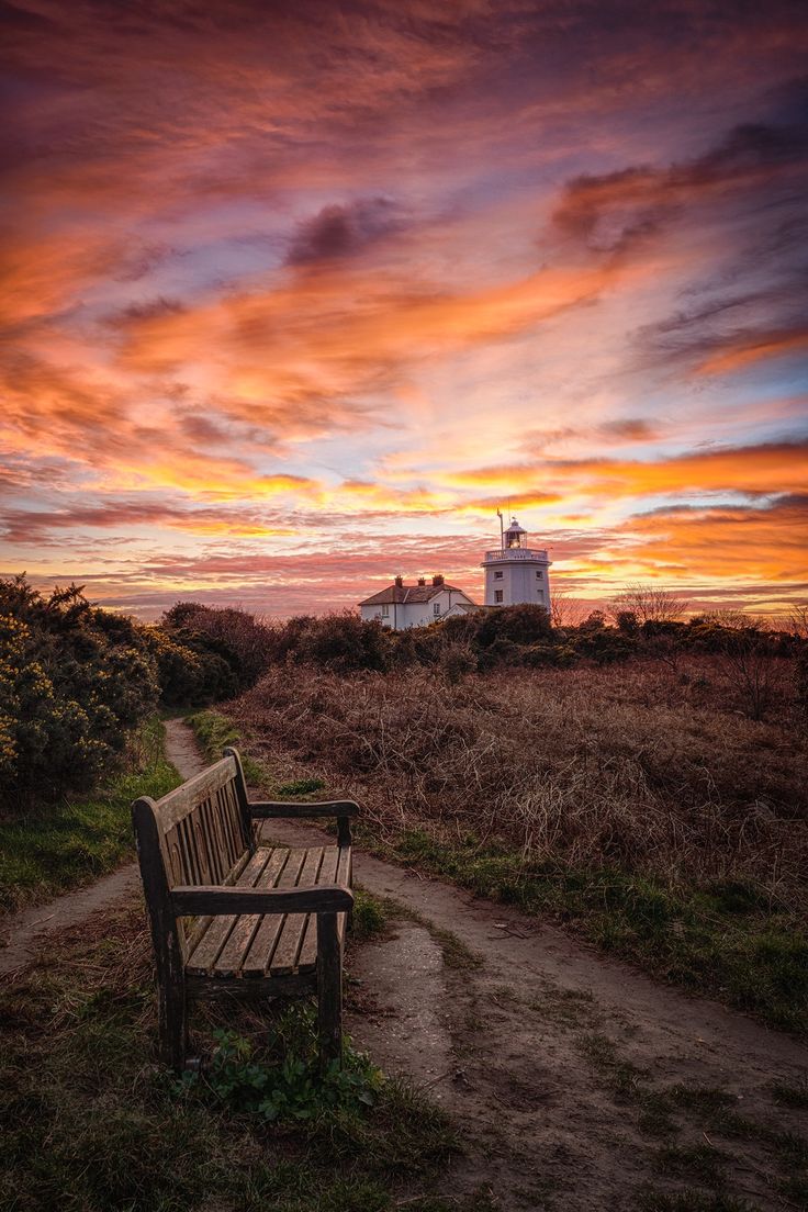 a wooden bench sitting on top of a grass covered field next to a lighthouse at sunset