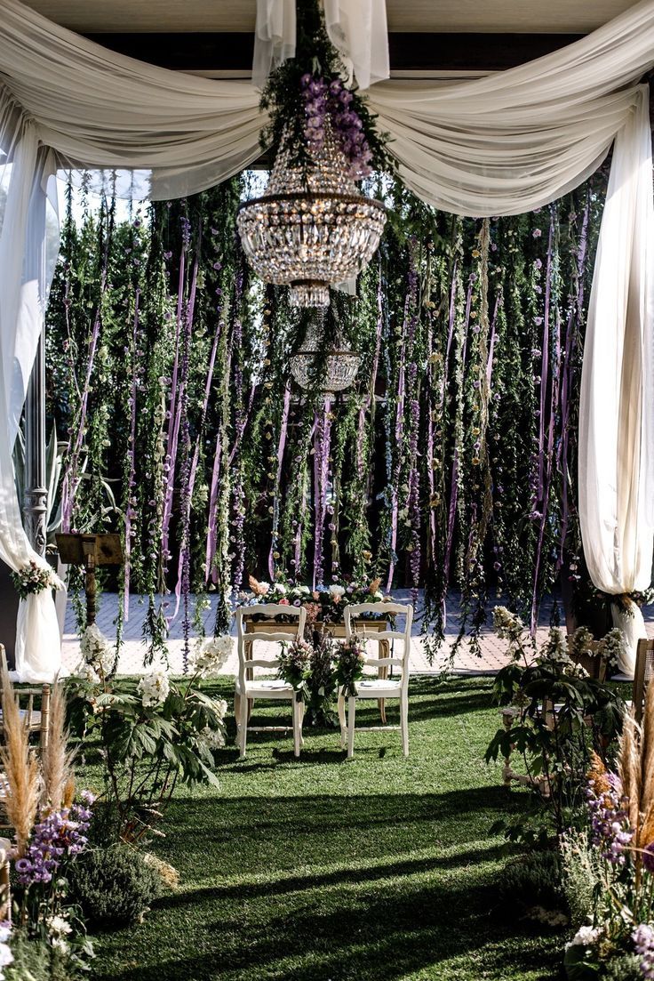 an outdoor wedding setup with purple flowers and greenery hanging from the ceiling, surrounded by white draping