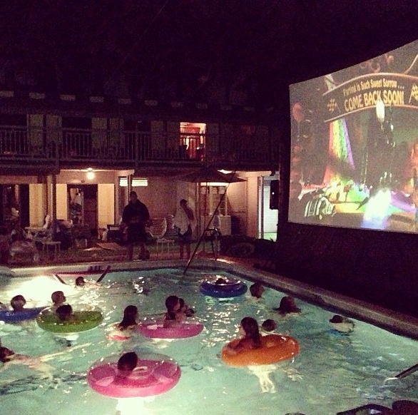 people are floating in the pool at night watching a movie on the wall behind them