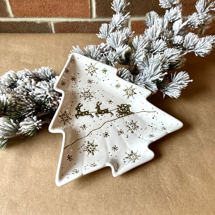 a white ceramic christmas tree dish next to some pine branches and snowflakes on a brown surface