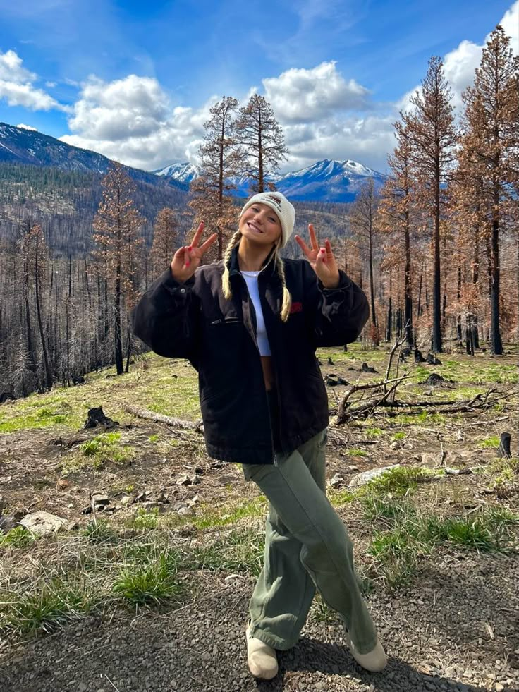 a woman standing in the middle of a forest with her hands up and two fingers out