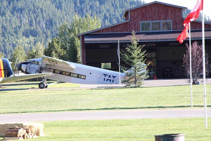 an airplane parked in front of a barn with two flags on the grass near it