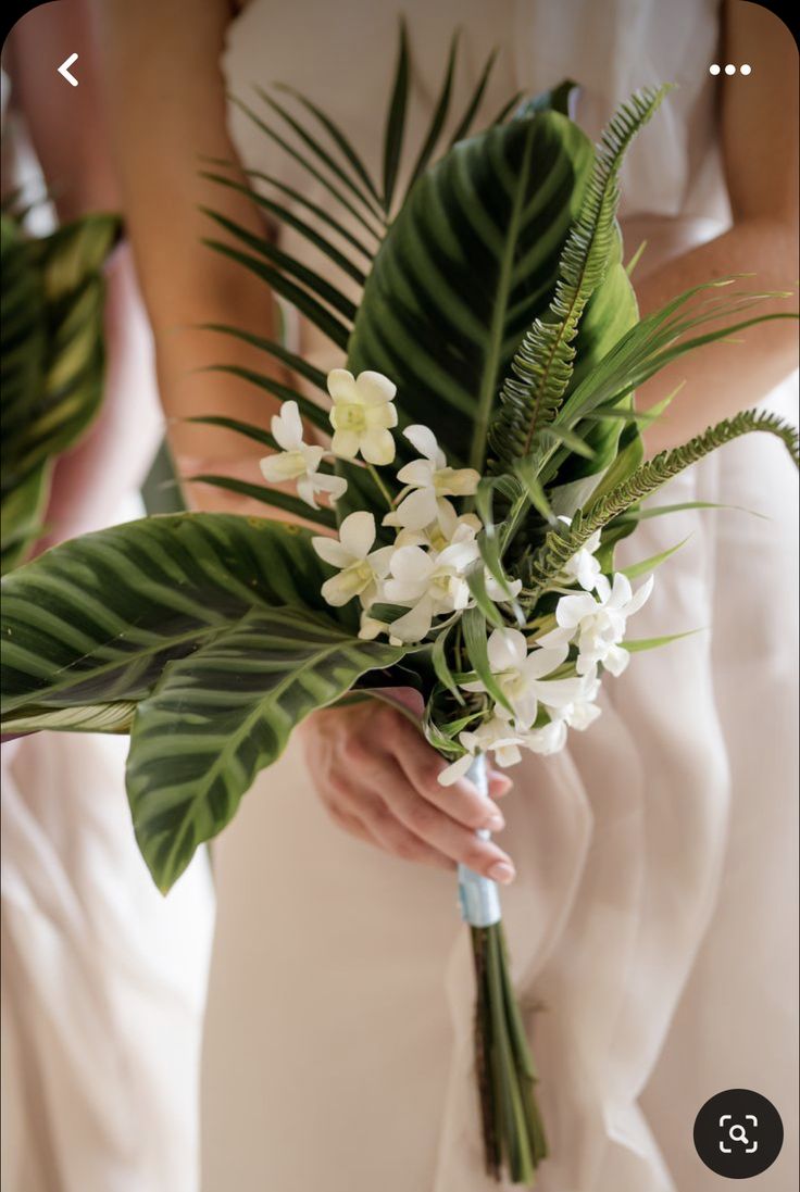 a bride holding a bouquet of white flowers and greenery in her hand with palm leaves