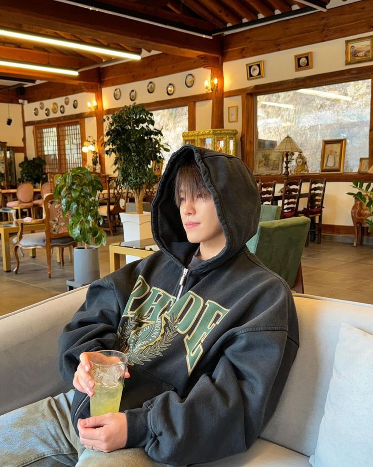 a young man sitting on top of a couch holding a drink in his hand and wearing a hoodie