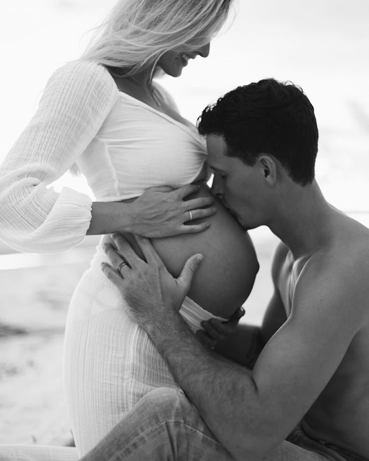 a pregnant man kisses his wife's belly on the beach in black and white