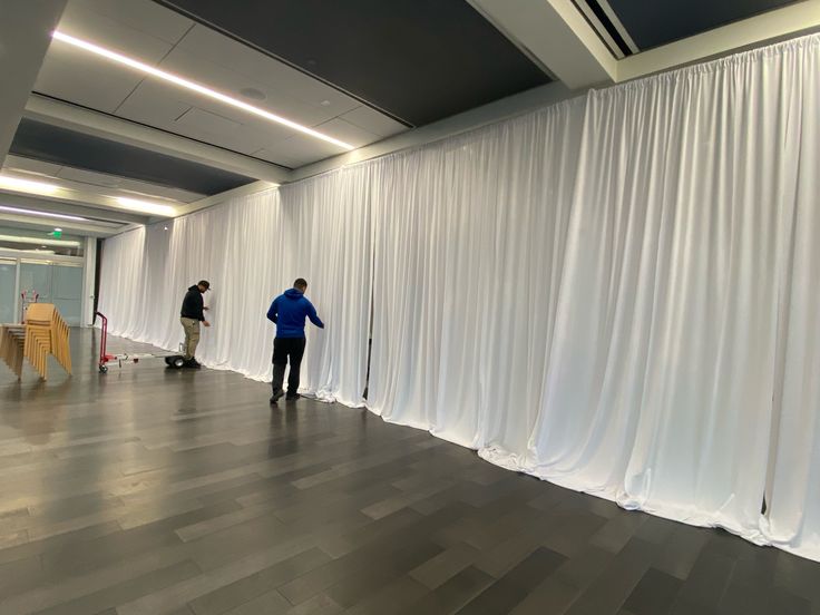 two men are standing in front of white drapes on the wall and one man is using a mop