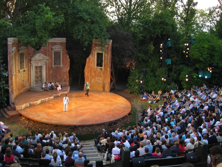 an outdoor stage with people sitting and standing on the steps in front of it, surrounded by trees