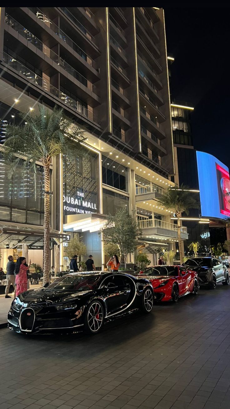 a group of exotic cars parked in front of a building at night with people walking by