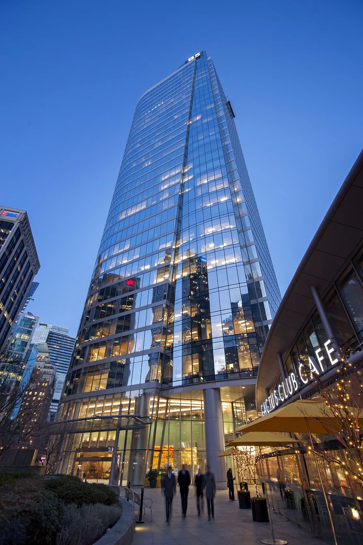 people are walking in front of a glass building at night with lights reflecting off the windows