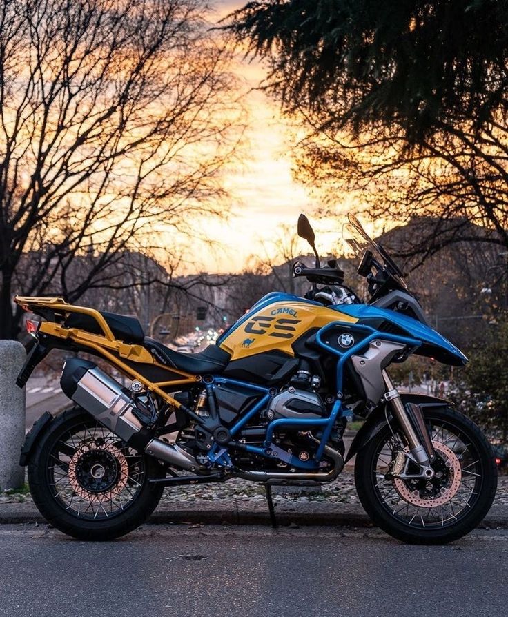 a blue and yellow motorcycle parked on the side of the road near trees at sunset