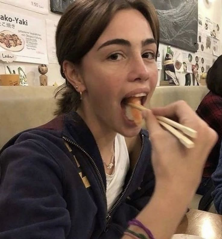 a woman sitting at a table with chopsticks in her mouth while eating food