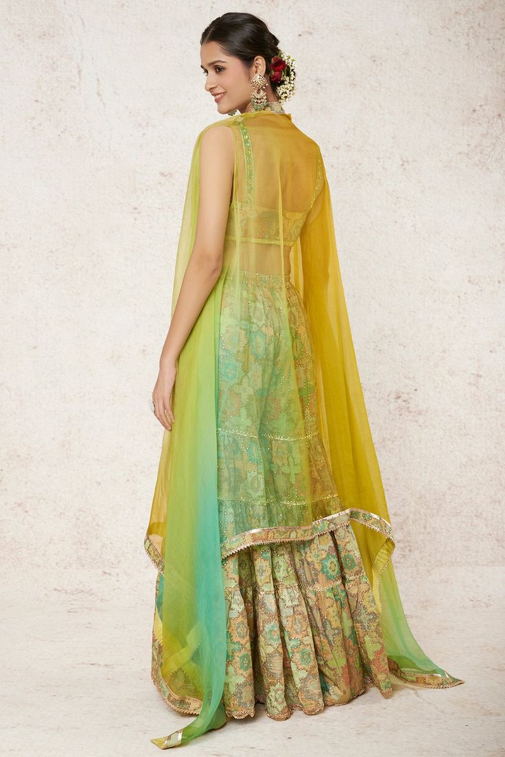 Editor's Note Celebrate with flair at mehndi and wedding parties in this youthful ensemble, tailored perfectly for you. The tiered sharara showcases a playful combination of lines and gota, adorned with whimsical designs and charming frill trim. The blouse finds its unique essence with an offset cape, creating a bold and perfect statement for the fun and adventurous spirit within you. Embrace this distinctive outfit and make unforgettable memories on your special occasions! Fabric: Tussar silk, Bollywood Style Ruffled Dresses For Navratri, Green Palazzo Set With Sheer Dupatta In Traditional Drape, Bollywood Style Sharara With Ruffles And Traditional Drape, Traditional Sharara With Ruffles For Navratri, Navratri Ruffled Sharara With Traditional Drape, Festive Anarkali Palazzo Set With Ruffles, Designer Ruffled Palazzo Set For Diwali, Festive Green Palazzo Set With Gota Work, Bollywood Style Ruffled Sharara For Navratri