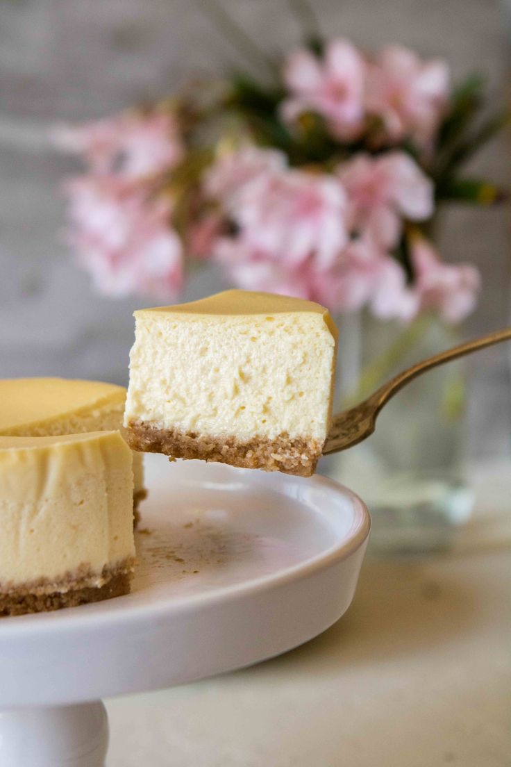 a piece of cheesecake on a white plate with a fork in it and some pink flowers