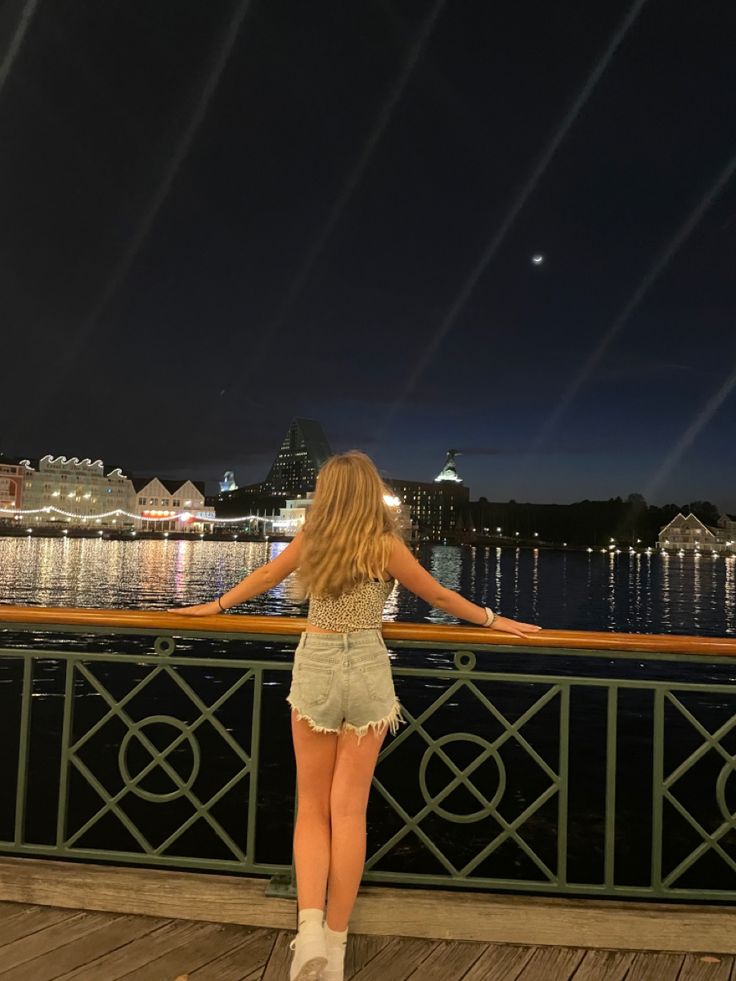 a woman standing on top of a wooden deck next to the water at night time