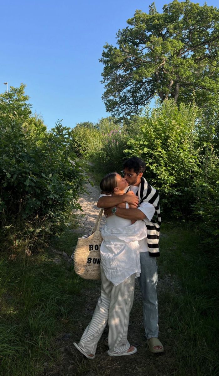 a man and woman kissing on the side of a dirt road in front of trees