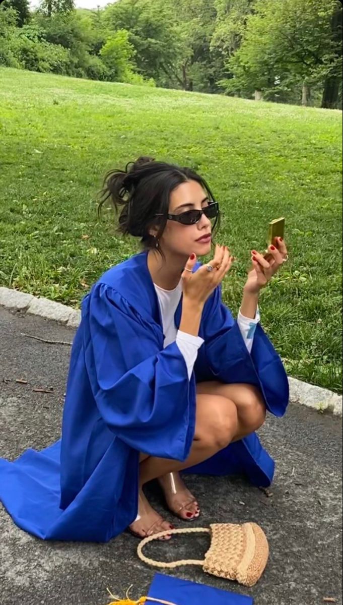 a woman sitting on the ground taking a photo with her cell phone wearing a blue graduation gown