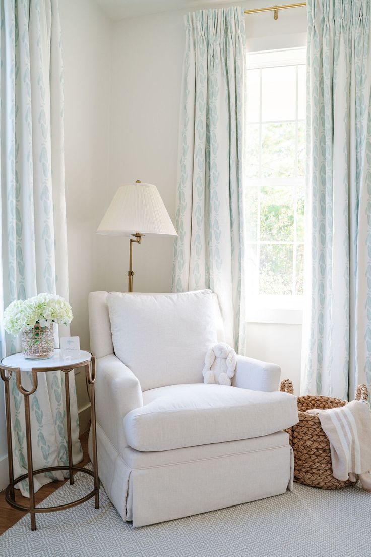 a white chair sitting in front of a window next to a table with a lamp on it