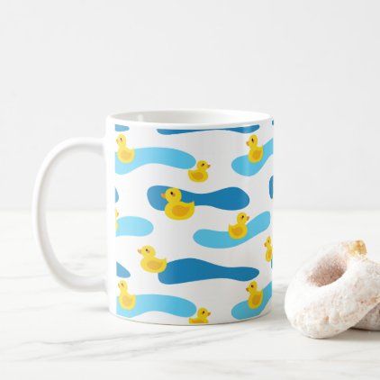 a white coffee mug with blue and yellow rubber ducks on it next to a donut