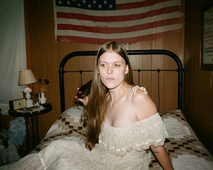 a woman sitting on top of a bed in a room with an american flag hanging above her head