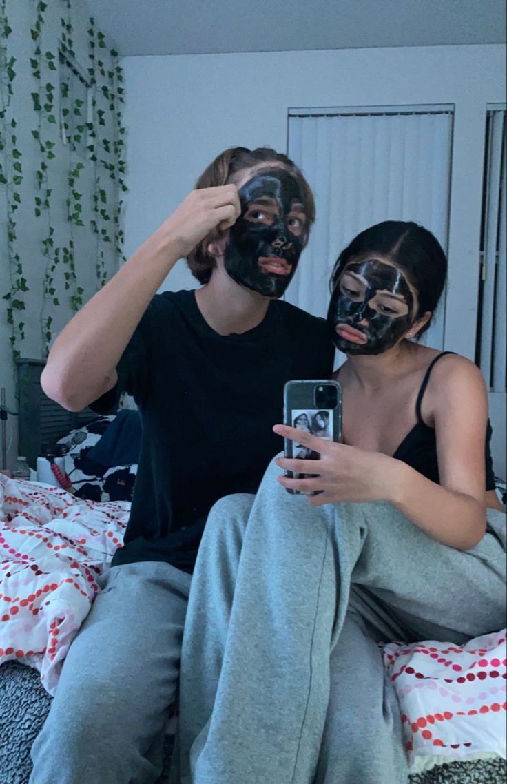 a man and woman sitting on a bed with their faces painted like masks looking at a cell phone