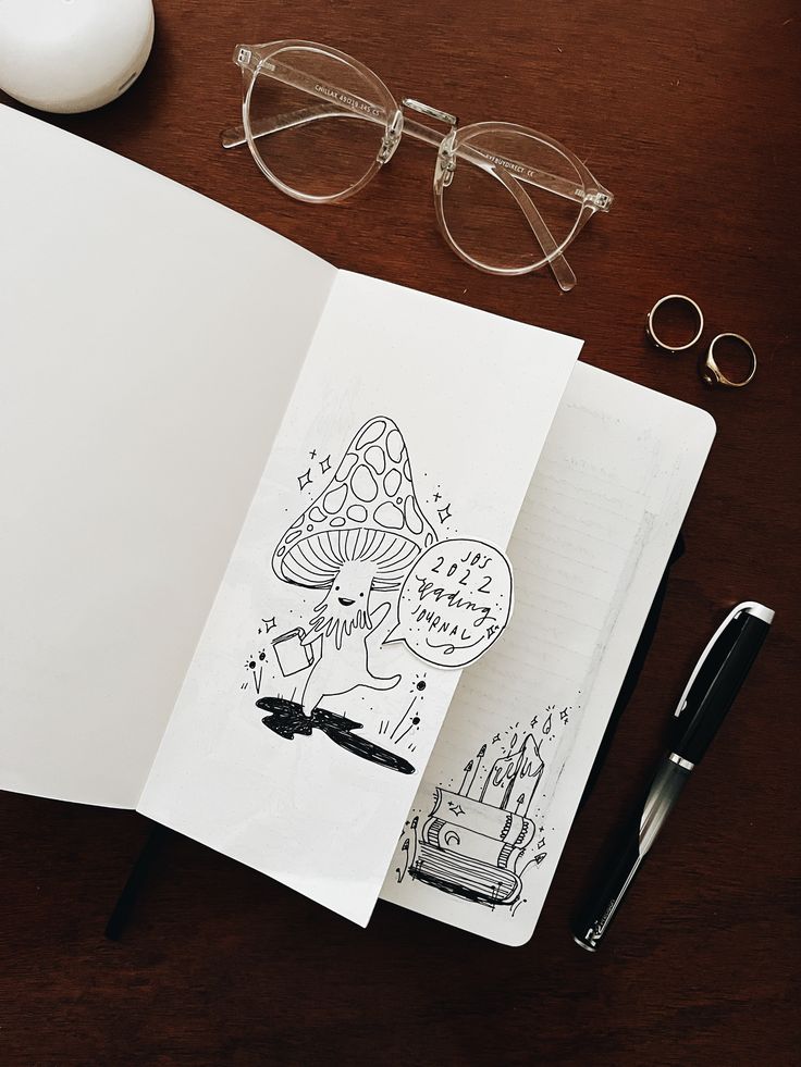 an open book with drawings on it next to a pair of glasses and a pen