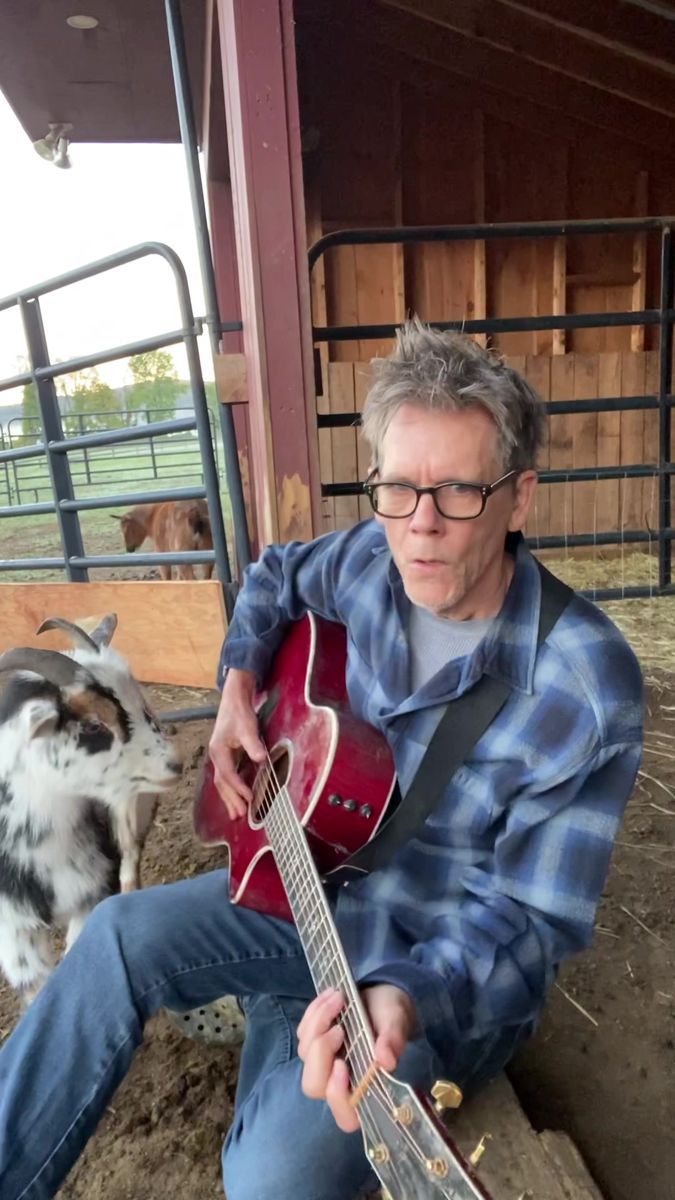 a man sitting on the ground playing an acoustic guitar next to a goat in a pen