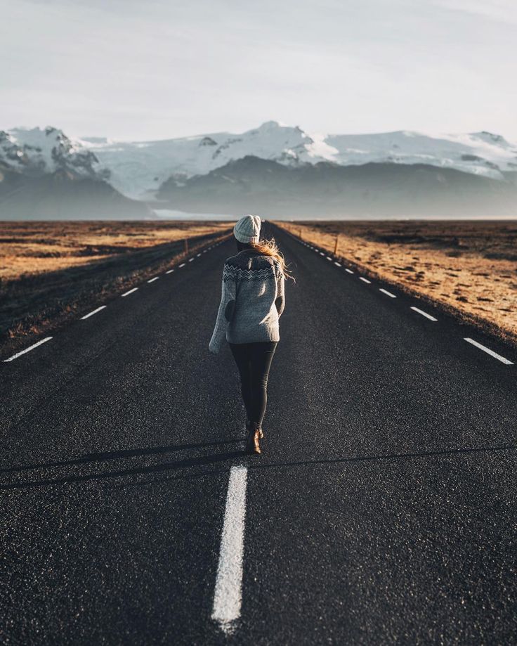 a woman walking down the middle of an empty road in front of snow capped mountains